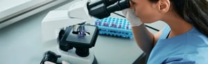 Labormitarbeiterin blickt durch ein Mikroskop zur Veranschaulichung von Post-Market-Surveillance-Best-Practices (PMS) nach MDR und IVDR.