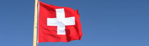 Schweizer Flagge auf einem Holzmasten weht vor einem verschneiten Alpengipfel unter blauem Himmel