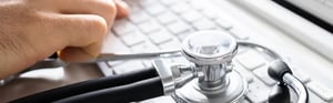 A stethoscope on a computer keyboard next to a person at work—a symbol of the role of medical experts in the clinical evaluation and regulatory compliance of medical devices.